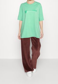 Osoba oblečená v volném zeleném tričku Karl Lagerfeld Jeans, hnědých širokých sametových kalhotách a bílých teniskách stojící na světlém pozadí.