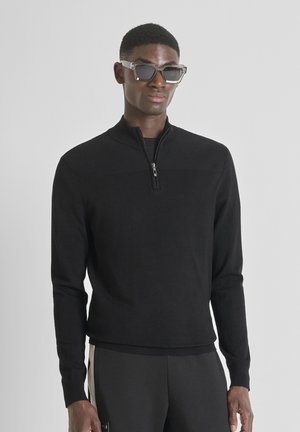 Sweat à zip noir avec des accents côtelés sur les épaules, porté sur une chemise noire, associé à des lunettes transparentes angulaires.
