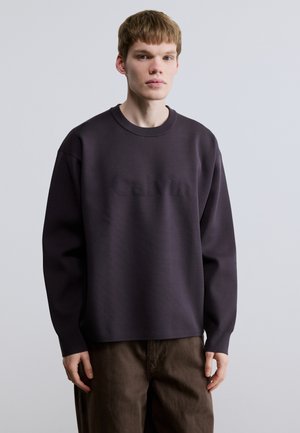 Sweatshirt gris foncé à col rond avec un logo Calvin texturé. Il présente des épaules larges et tombantes ainsi qu'une coupe décontractée, fabriqué dans un tissu doux.