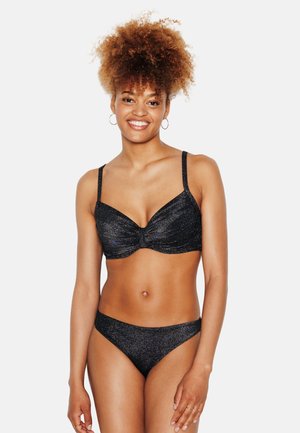 Donna sorridente con capelli ricci che indossa un top bikini nero scintillante e slip coordinati, in piedi contro uno sfondo bianco semplice.