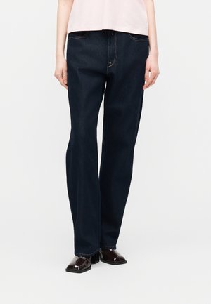 Person wearing dark blue straight-leg jeans, a light pink top, and black square-toe heeled shoes, standing against a plain background.