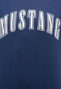 Mörkblå bomull t-shirt med stora vita bokstäver som stavar "MUSTANG" i en böjd, fet stil över bröstet. Slät textur, avslappnad design.