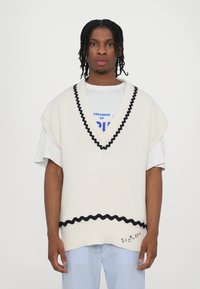 Top in maglia color crema con un bordo ondulato nero, indossato su una t-shirt bianca con testo multicolore, abbinato a pantaloni a righe blu.