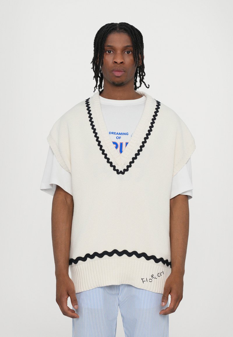 Top in maglia color crema con un bordo ondulato nero, indossato su una t-shirt bianca con testo multicolore, abbinato a pantaloni a righe blu.