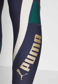 Leggings sportivi blu navy con pannelli verdi e crema, caratterizzati da un prominente logo dorato "PUMA" e dettagli di cucitura eleganti.