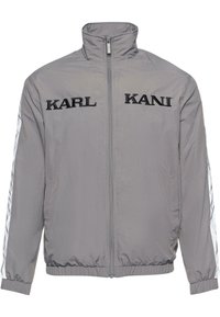 Grey zip-up jacket with high collar, featuring black embroidered "KARL KANI" logo on the front, and white striped accents on the sleeves.