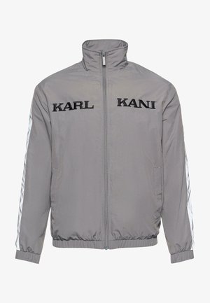 Chaqueta gris con cremallera y alto cuello, que cuenta con el logo bordado en negro "KARL KANI" en la parte delantera y acentos de rayas blancas en las mangas.
