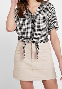 Kvinna i en svart-och-vit rutmönstrad skjorta med knappar, knuten i midjan, i kombination med en beige mini kjol. Avslappnad och stilren outfit.