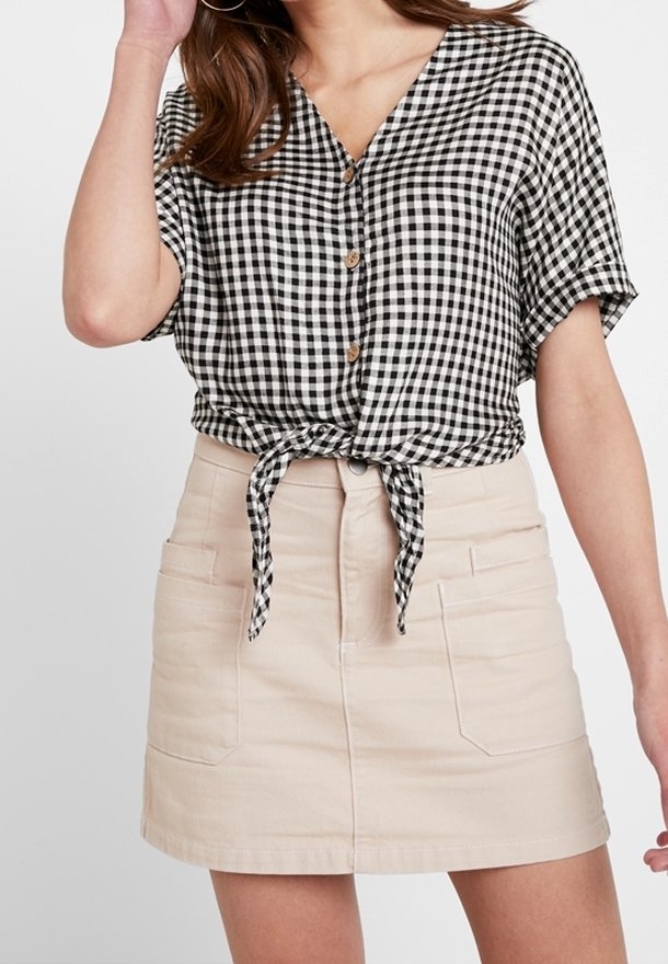 Kvinna i en svart-och-vit rutmönstrad skjorta med knappar, knuten i midjan, i kombination med en beige mini kjol. Avslappnad och stilren outfit.