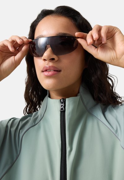 Person, der justerer store, mørke solbriller, iført en lys grøn lynlåstrøje med høj krave mod en ensfarvet baggrund.