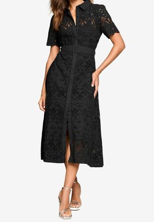 Femme portant une robe midi en dentelle noire avec des manches courtes, un col, des boutons, une ceinture assortie et des sandales à talons hauts argentées.