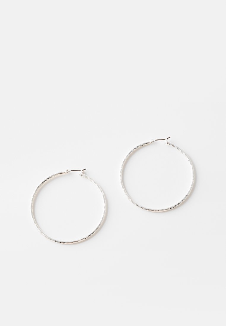 Dos pendientes de aro retorcidos, delgados y plateados con un pequeño broche sobre un fondo blanco liso.