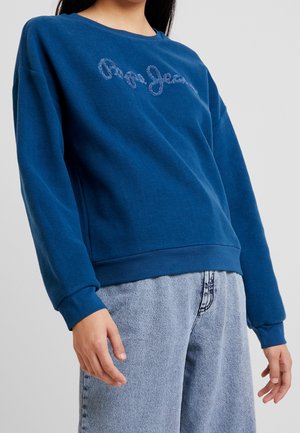 Person wearing a blue long-sleeve sweater with embroidered text "Pepe Jeans" and light blue denim jeans against a plain background.