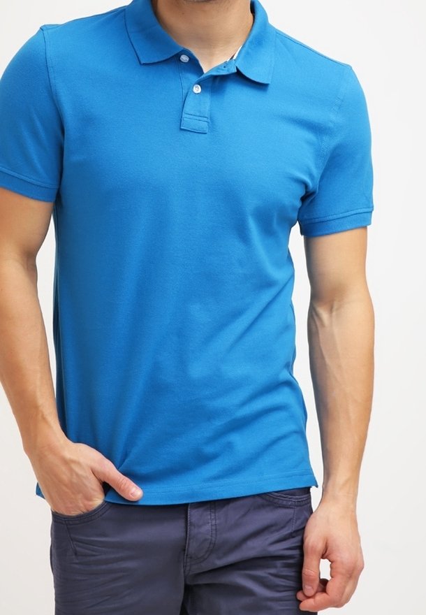Homme portant un polo bleu vif à manches courtes et un pantalon bleu marine foncé, debout avec une main dans la poche devant un fond uni.