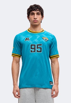 Jeune homme aux cheveux courts et foncés, portant un maillot de sport bleu sarcelle des Jacksonville Jaguars avec le numéro 95 et un short gris sur un fond uni.