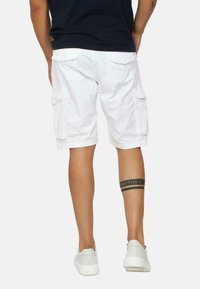 Pantaloni cargo bianchi con tasche laterali, materiale in cotone e vestibilità rilassata. Un braccialetto tatuato nero sulla gamba sinistra riporta il testo "FRATELLO."