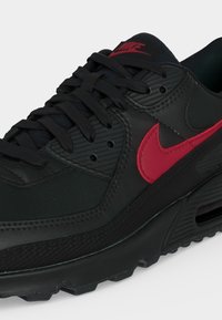 Μαύρο αθλητικό παπούτσι Nike Air Max με μαύρα κορδόνια και κόκκινο λογότυπο Swoosh στο πλάι και στη γλώσσα, σε λευκό φόντο.