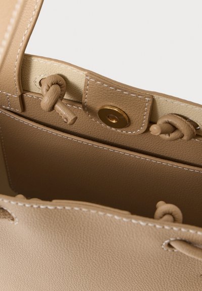 Μπεζ δερμάτινη τσάντα tote με υφή, με κόμπους ως διακοσμητικά και κούμπωμα με μαγνητικό κλιπ. Το εσωτερικό είναι επενδυμένο σε ανοιχτότερη απόχρωση.