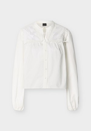 Blouse blanche à manches longues avec boutons sur le devant et détails en dentelle florale brodée sur la partie supérieure de la poitrine et les épaules.