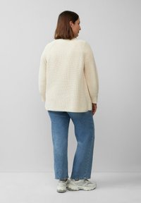 Cremefarbener Strickpullover mit strukturiertem Muster, langen Ärmeln und lockerer Passform, kombiniert mit hellblauen geraden Jeans und grauen Sneakers.