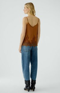 Vrouw die zijwaarts kijkt, gekleed in een bruin camisole met kant, een blauwe spijkerbroek, zwarte haklaarzen en pareloorbellen, tegen een effen achtergrond.