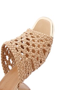 Beige geweven slip-sandaal met een gestructureerde bovenkant, uitgevoerd met een gevlochten design en een gladde zool. Vierkante hak en subtiele randafwerking.