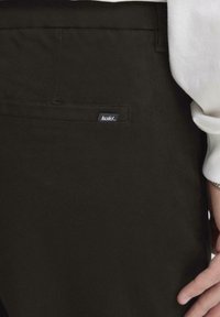 Los pantalones marrón oscuro tienen un bolsillo trasero con una pequeña etiqueta negra. La tela parece suave y resistente, con un diseño estructurado.