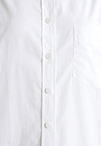 Camicia bianca con bottoni, caratterizzata da una placca con bottoni completa, un'unica tasca sul petto, una texture liscia e un design semplice.