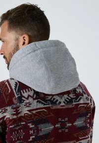 Hættetrøje med grå hætte, med en rød mønstret krop, der viser stammemotiver i beige og marineblå accenter.