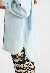 Lichtblauw denim overhemd met een afgeronde zoom en lange mouwen, gecombineerd met zwarte en beige broeken met een patroon van golvende ontwerpen.
