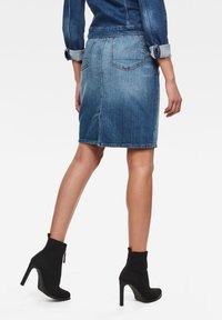 Jupe en denim de bleu clair à moyen avec une coupe droite, des poches arrière et une fente à l'arrière. Portée avec des bottines noires à talons hauts.