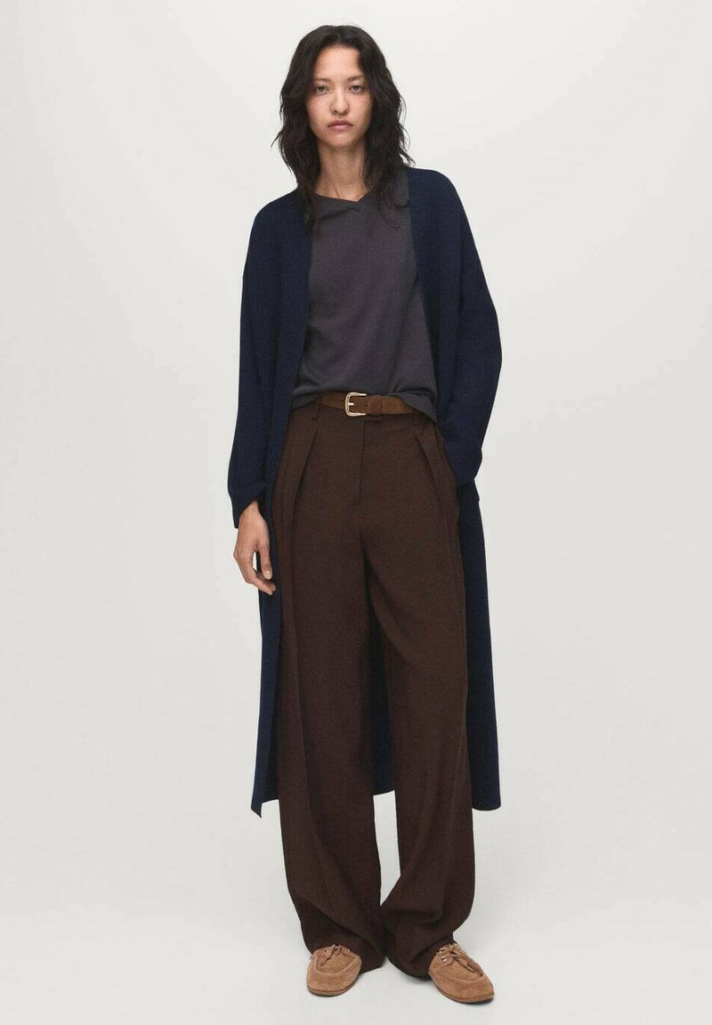 Vrouw in een donkergblauwe lange cardigan, grijze top, bruine wijde broek met riem en tan moccasinschoenen, staand tegen een effen achtergrond.