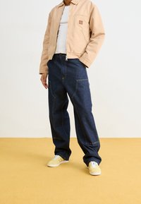 Giacca di jeans color sabbia con tasca frontale, abbinata a jeans blu larghi con cuciture gialle e sneakers giallo chiaro.