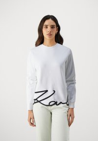 Bílá mikina s dlouhými rukávy a kulatým výstřihem. Na lemu se nachází výrazné černé kurzivou napsané logo "Karl". Hladká textura.