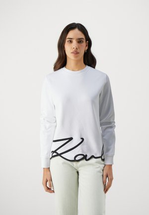 White sweatshirt with long sleeves and a crew neckline. Features a prominent black cursive "Karl" logo at the hem. Smooth texture.