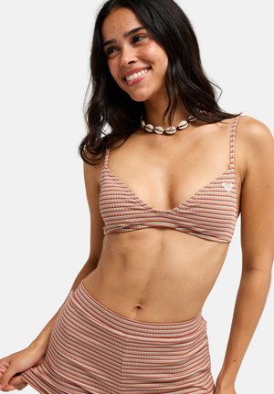 Młoda kobieta uśmiechająca się, ubrana w paskowany top od bikini i dopasowane majtki, z białym naszyjnikiem z muszli na jednolitym tle.