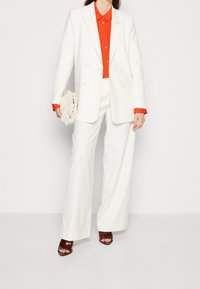 Vit dubbelknäppt kavaj, i kombination med en orange skjorta och matchande vida byxor. Accessoarer med en beige clutchn och bruna klacksandaler.