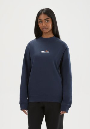 Frau mit langen schwarzen Haaren, die einen dunkelblauen ellesse Pullover und schwarze Hosen trägt, steht vor einem einfachen hellen Hintergrund.