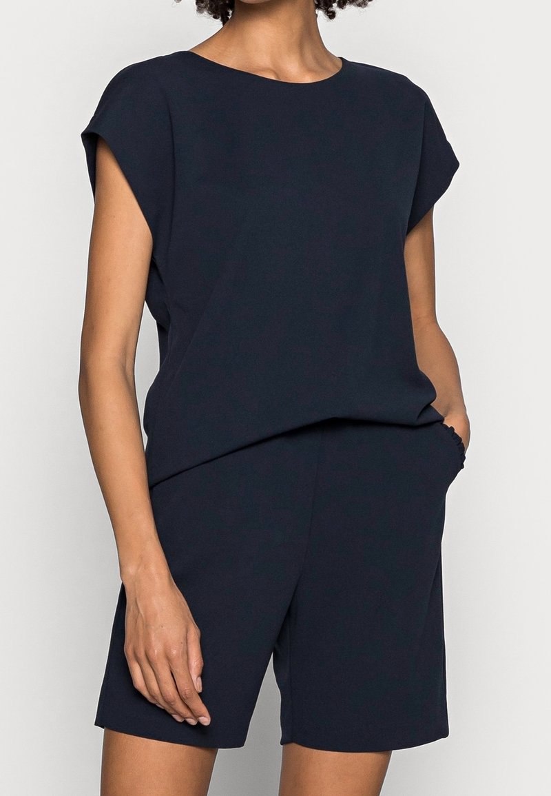 Osoba w bezrękawnikowej czarnej bluzce i pasujących czarnych szortach, z jedną ręką w kieszeni, na jednolitym tle.