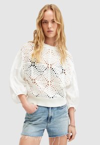 Jersey blanco con un intrincado patrón de cortes, mangas abullonadas de tela blanca, combinado con pantalones cortos de mezclilla azul claro, con dobladillo desflecado.