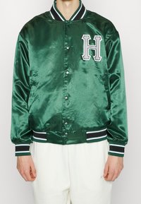 Grüne Satin-Bomberjacke mit einem weißen "H"-Patch, gerippten schwarzen und weißen Bündchen sowie Druckknopfverschluss. Glatte Textur, sportliches Design.