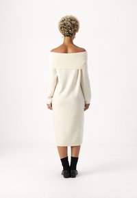Φόρεμα με ανοιχτούς ώμους, μακριά μανίκια, σε απαλό κρεμ ύφασμα, μήκους μέχρι το γόνατο, με ριχτό λαιμόκοψη και μανσέτες, σε συνδυασμό με μαύρες κάλτσες μέχρι τους αστράγαλους και παπούτσια.
