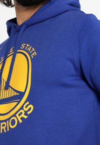 Sudadera azul hecha de una mezcla de algodón, con un gran logo gráfico amarillo que presenta un puente y el texto "WARRIORS" en la parte delantera.