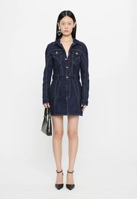 Denim dress in deep blue with contrast stitching, button closure, shirt collar, belted waist, and long sleeves. Paired with black heels and bag.