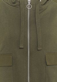 Primo piano di una felpa verde oliva frontale che mostra la cerniera argentata con tirante rotondo e i lacci coordinati sopra due tasche a pattina.