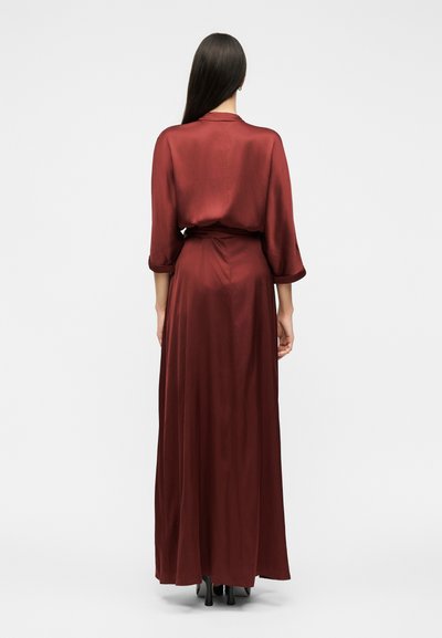Femme aux longs cheveux foncés porte une robe en satin bordeaux longue jusqu'au sol avec des manches trois-quarts, debout de dos sur un fond uni.