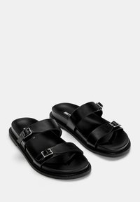 Sandales en cuir noir avec deux larges sangles et boucles en argent. Semelle intérieure rembourrée et semelle plate en caoutchouc pour un confort optimal. Design minimaliste.