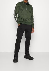 Maglione zip-up verde con il logo Nike sul petto, con accenti neri lungo le maniche, abbinato a pantaloni neri e sneaker nere.