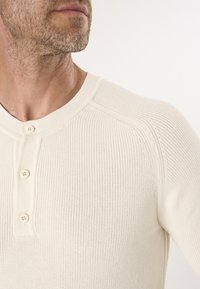 Cremefarbenes Strickshirt mit gerippter Textur, rundem Ausschnitt und einer Knopfleiste mit zwei Knöpfen auf der Vorderseite.