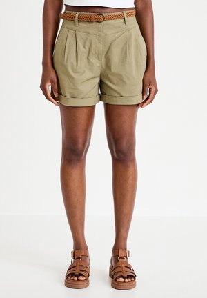 Benedenbenen met opgerolde beige korte broek, gevlochten riem en bruine sandalen met bandjes tegen een effen witte achtergrond.